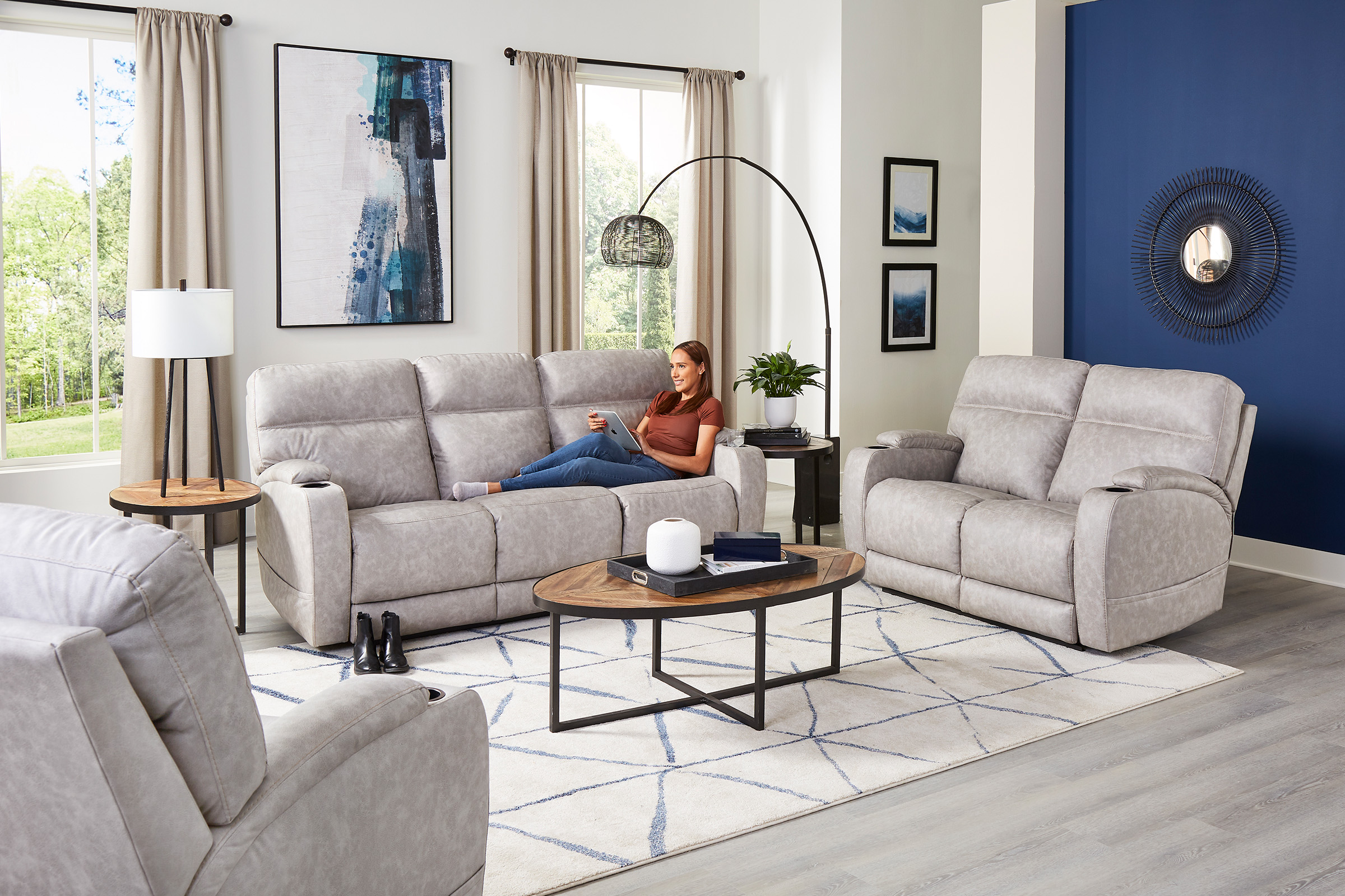 A woman lays down on a light grey upholstered couch, looking at her tablet. A matching loveseat and chair sit next to the couch.