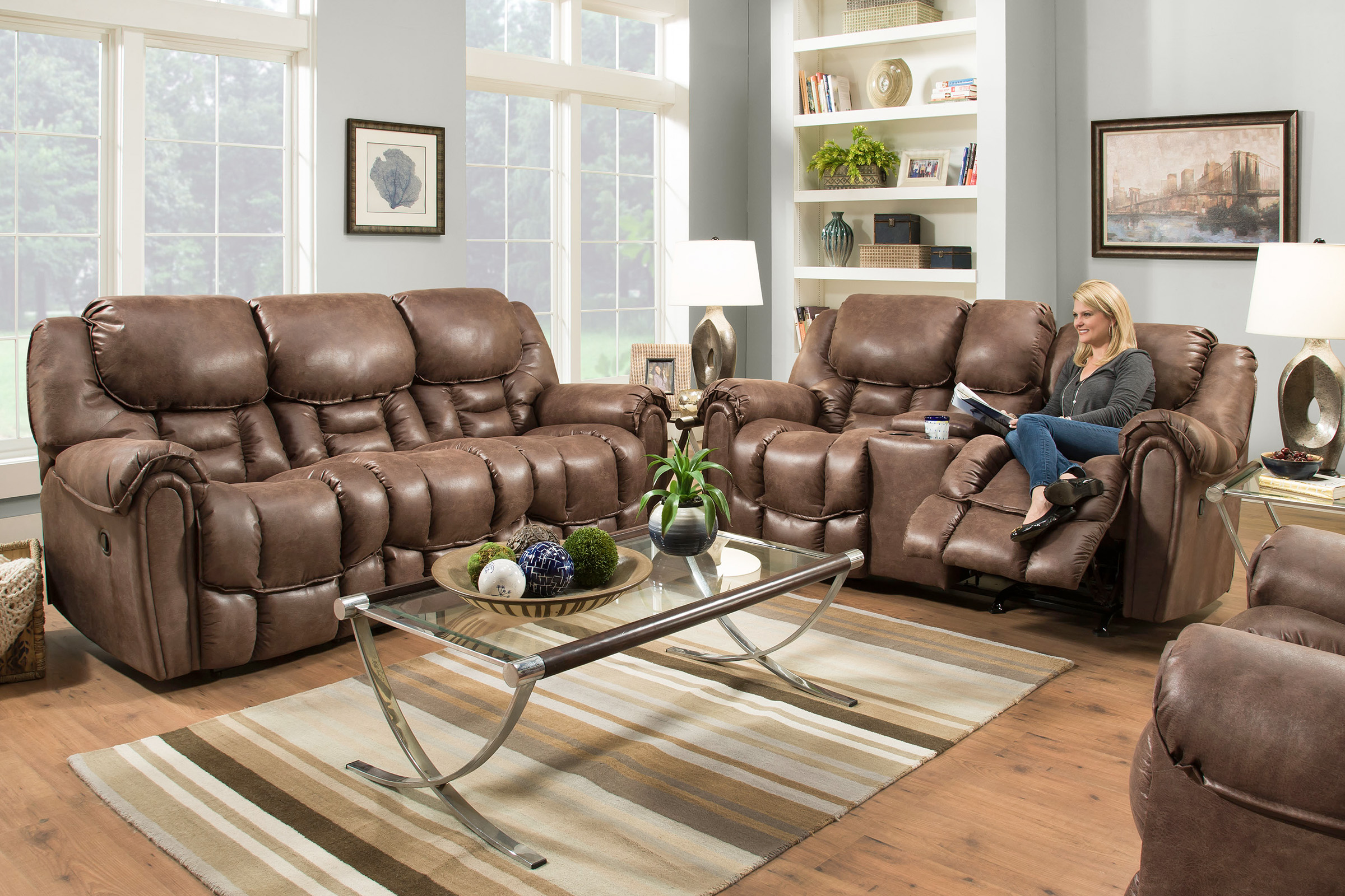 A lady sits on a cushioned, brown leather loveseat with the footrest raised up. A matching sofa sits next to it.